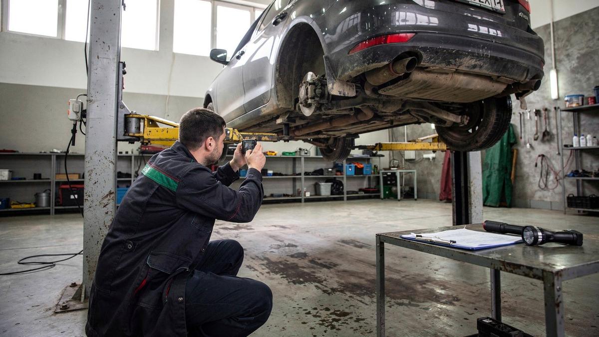 Independent inspector photographing the underside of a dark sedan raised on a lift in a Kyiv workshop